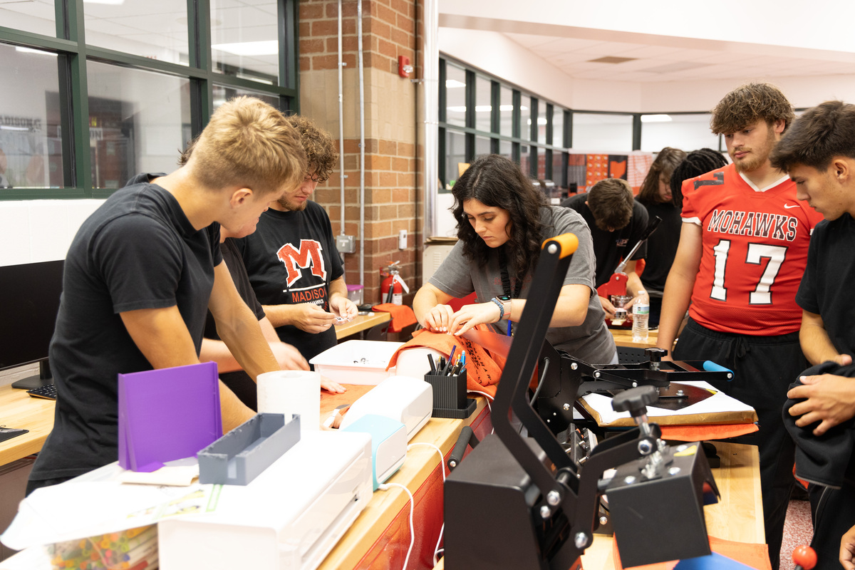 A teacher shows multiple students standing around equipment how to add text to a towel.